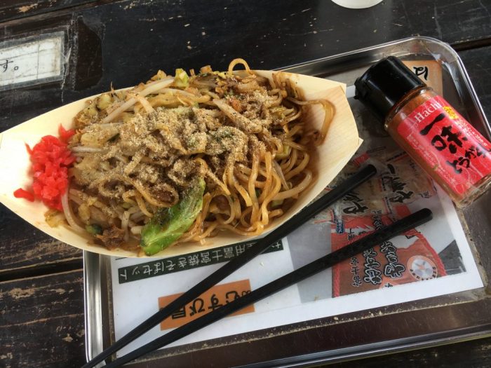 むすび屋の焼きそばを撮影。銀色のトレイの上に紙で出来た皿に盛られた焼きそば、一味、黒いピラスチックの箸がある。